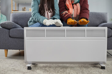 Cropped photo of a frozen couple sitting on the sofa in the living room in winter outerwear and gloves at home and trying to warm their hands on electric heater. Heating problems concept. - obrazy, fototapety, plakaty
