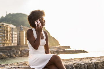 Young afro woman sitting on a promenade talking with mobile - obrazy, fototapety, plakaty