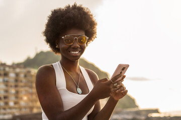 Stylish african woman using phone outdoors during sunset