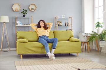 Happy girl relaxing on sofa after work at home. Attractive dreaming young woman in casual clothes sitting on comfortable couch with hands behind head enjoying free time, lazy weekend time