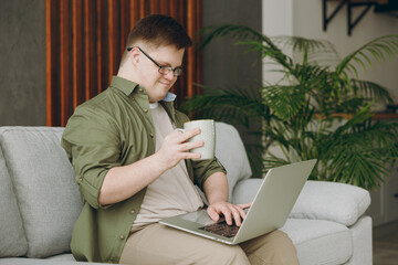 Young IT man with down syndrome wear casual clothes work hold use laptop pc computer sits on grey sofa couch staying at home flat rest spend free time in living room Genetic disease world day concept