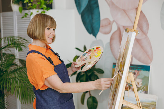 Side profile view elderly fun confident artist woman 50 years old wears casual clothes stand near easel with painting artwork paint spend free spare time in living room indoor. Leisure hobby concept