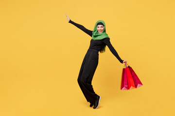 Full body young asian muslim woman wears green hijab abaya clothes hold shopping package bags lean back stand on toes isolated on plain yellow background. Black Friday sale buy day, uae islam concept.