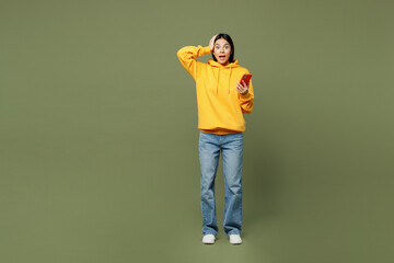 Full body young excited surprised overjoyed Latin woman wears yellow hoody casual clothes hold head use mobile cell phone isolated on plain pastel green background studio portrait. Lifestyle concept.