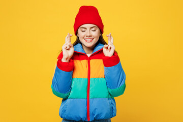 Young woman she wears padded windbreaker jacket red hat casual clothes waiting for special moment, keeping fingers crossed, making wish isolated on plain yellow background studio. Lifestyle concept.