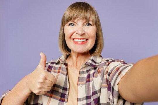 Close up elderly woman 50s years old wearing beige t-shirt shirt casual clothes doing selfie shot pov on mobile cell phone show thumb up isolated on plain pastel purple background. Lifestyle concept.