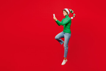 Full body young woman wear green turtleneck Santa hat posing jump high hold in hand use mobile cell phone isolated on plain red background. Happy New Year 2024 celebration Christmas holiday concept.