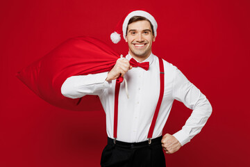 Fototapeta na wymiar Merry young smiling man wear white shirt Santa hat posing hold bag with present gifts look camera isolated on plain red background studio portrait Happy New Year Christmas celebration holiday concept