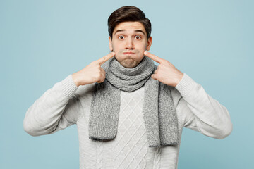Young sick man in gray sweater scarf point fingers on blowing puffing cheeks isolated on plain blue background studio portrait. Healthy lifestyle disease virus treatment cold season recovery concept.