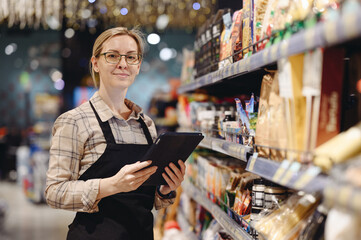 Young seller woman wears uniform hold digital tablet pc computer check aisle arrange for shopping at supermaket store grocery shop buying choose products inside hypermarket. Purchasing food concept.