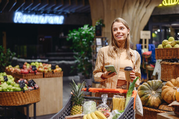 Young customer woman wear casual clothes using mobile cell phone drinking coffee shopping with trolley cart at supermaket store grocery shop buy choose products in hypermarket Purchasing food concept.