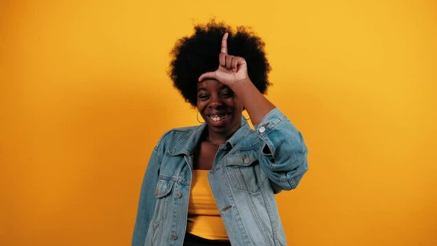 Young African American woman showing loser hand gesture while posing against yellow studio background. Emotions. People. Lifestyle concept