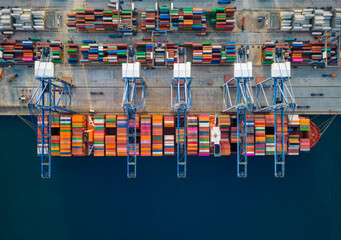Aerial top down view of a large container cargo ship being unloaded by cranes and trucks in a commercial harbour