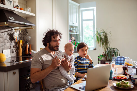 Dad multitasking with baby while son enjoys breakfast and laptop