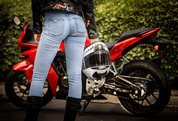 Close up of woman in blue jeans holding a motorcycle helmut with a street motorcycle in the background 001 - obrazy, fototapety, plakaty