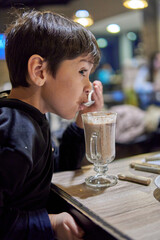 Latino brunette little boy in profile in a bar drinking hot chocolate milk with a spoon. Background with blurred lights.