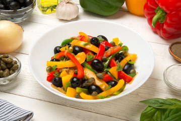 Roasted bell peppers with olives and capers in a salad bowl with ingredients on a white rustic table. - 671764595