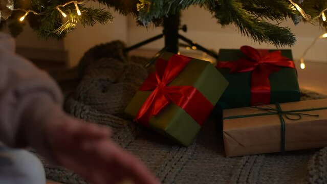 Close-up hands of unrecognizable woman carefully putting wrapped Christmas gift boxes with presents under xmas tree in room with festive interior. Happy female person making surprise on xmas eve