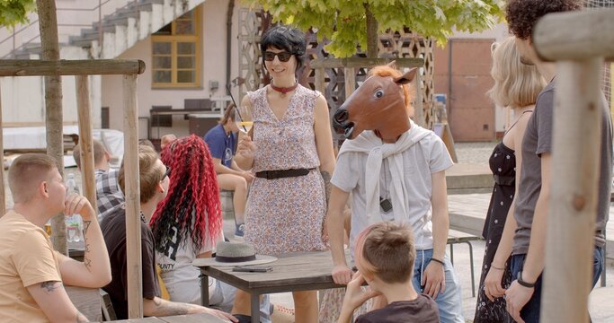 Young woman holding glass of non-alcoholic beverage joins group of young people. Her appearance is unconventional for teacher, reflecting sense of freedom in self-expression. Man wearing a horse mask