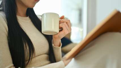 Carefree young woman in warm sweater drinking hot tea and reading book
