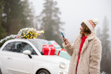 Woman using phone while traveling by car decorated with presents and Christmas tree in mountains. Girl having fun celebrating winter holidays on nature