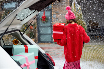 Woman in red sweater and hat carries wrapped Christmas tree and a present to her house on snow fall, View from behind. Concept of preparing for winter holidays