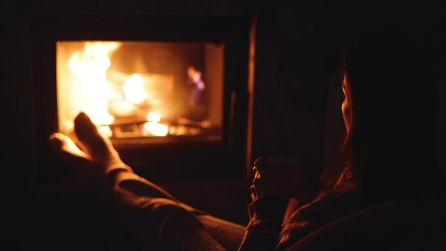 Warming up by the fire, cozy night in the cabin by the fireplace, fireplace burns in the Scandinavian cottage chalet house, burning fire with charcoal and firewood, young woman with cup of tea drink