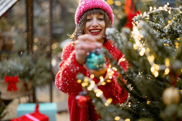 Young happy woman in red decorates lush Christmas tree with festive ballls and garland at backyard of her house on snow fall, preparing for a winter holidays