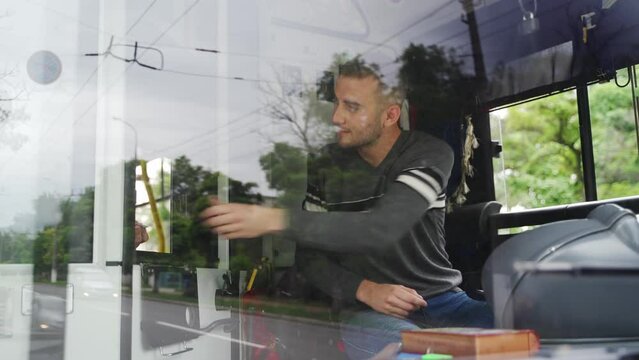 Polite professional transport operator sits on driver's seat, takes fare and gives change and ticket through small window. Young driver in casual clothes receives travel cost from passanger