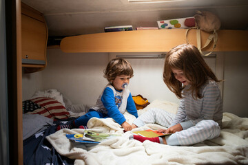 Two caucasian children, brother and sister, reading books in pajamas on a campervan bed during a...