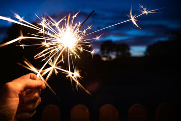 Hand holding a sparkler, or bengal fire stick, burning in outdoor setting at blue hour dusk. Holidays or magic background or wallpaper.