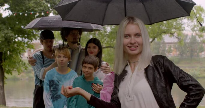 A group of joys pose under an umbrella in rainy weather. Blonde woman touches an umbrella, smiles. Positive mood in a friendly company.