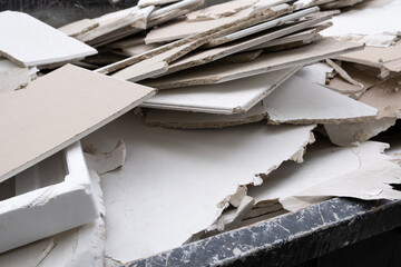 A huge overloaded dumpster waste container, overflowing with construction waste, drywall, and other rubble, stands near a construction site. The bin is filled to the brim with debris and garbage