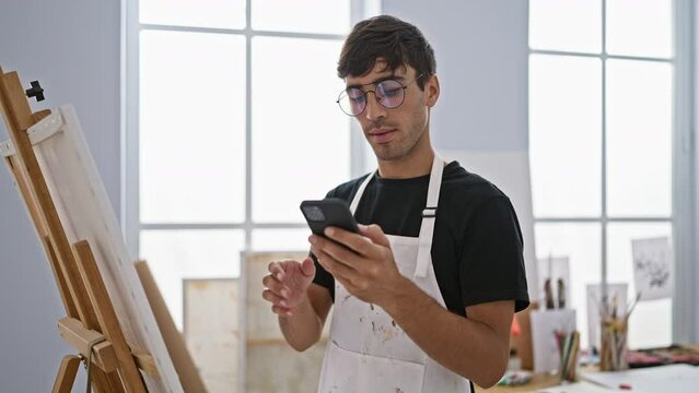Handsome young hispanic man artist immersed in creativity, seriously concentrating while texting on smartphone amidst paintbrushes and canvas in the heart of art studio
