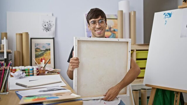 Smiling, confident young hispanic man draws passionately at art studio, a portrait of a handsome, smiling artist with paintbrushes in hand, basking in the warmth of creativity.