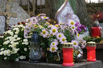 Grave with glass candles and flowers in pots among dry autumn leaves in cemetery on November day