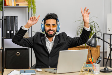 Happy relaxed overjoyed Indian businessman working on laptop computer at home office wearing headphones listening favorite energetic disco music and dancing. Freelancer man relaxing, taking a break