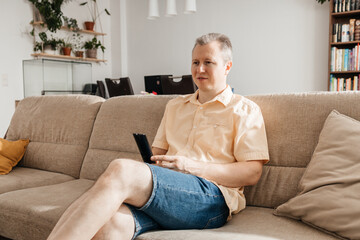 A man watches TV while sitting on the sofa in the living room on his day off and holds the TV remote control in his hands.