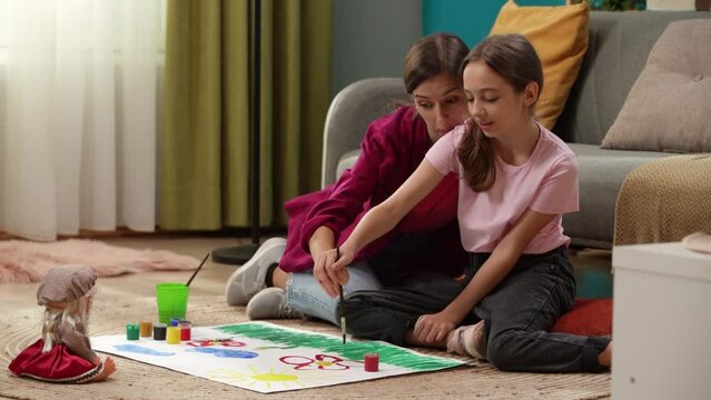 In a close up shot, a woman and a child sit on the floor and draw on paper together. They are communicating, spending time together. There is a sofa next to it, toys are scattered on the floor