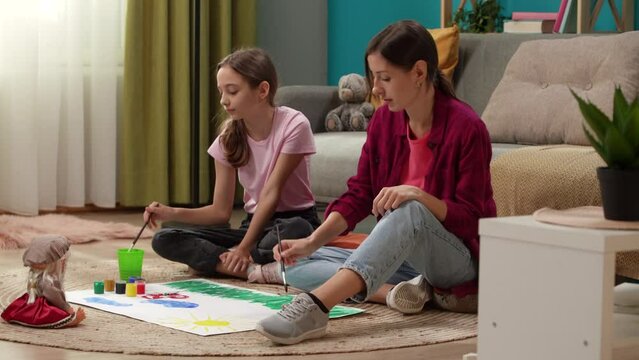 In a close up shot, a woman and a child sit on the floor and draw on paper together. They are communicating, spending time together. There is a sofa next to it, toys are scattered on the floor