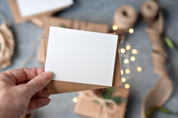 Hand holding blank paper card mockup and envelope over blurred table background with Christmas...