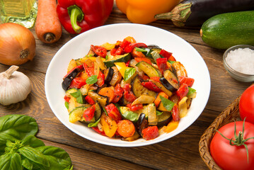 Sautéed  eggplant zucchini carrots and tomatoes in a salad bowl on a rustic wooden table with ingredients. - 671844501