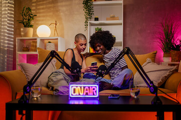 Two diverse women using phone and recording internet podcast in a studio