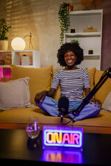 African woman sitting on a yellow sofa in home studio and recording podcast