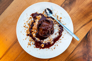 Delicious ice cream scoop on plate close-up with melted chocolate dressing and hazelnuts on a rustic wooden table