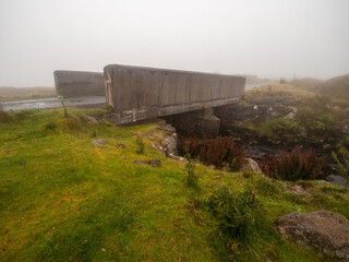 Fototapeta na wymiar Small simple concrete bridge for one car over a river or creek. Fog in the background. Driving in dangerous conditions due to poor visibility. Connemara, Ireland. Rural country side area.