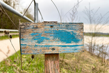 Wooden sign with peeling paint. A sign with old blue paint is embedded in the ground near the river and garden.