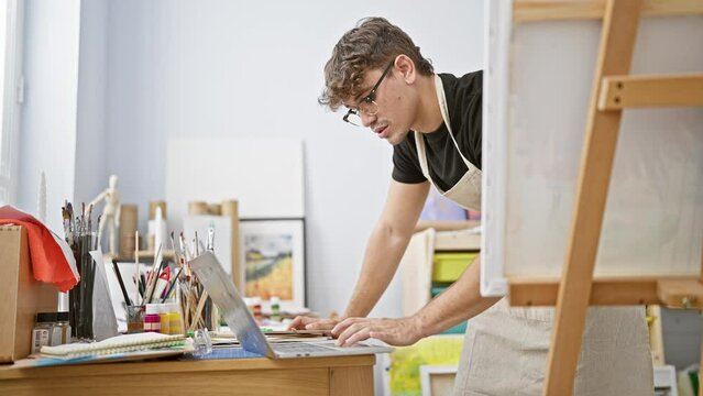 Attractive young hispanic man, a serious artist, standing by the easel in a well-lit art studio, passionately drawing on his laptop, immersed in the online creative sphere