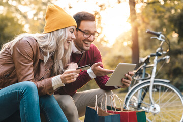 Cheerful young couple shopping online with credit card on a tablet in the city park.