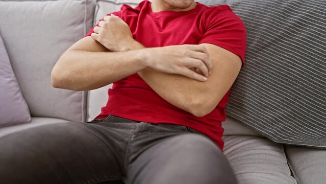 Portrait of a young, handsome hispanic man sitting on sofa at home, scratching itchy arms - a possible allergic reaction causing skin irritation and discomfort.